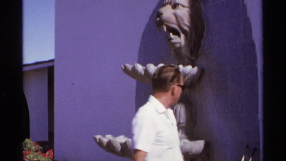 A male tourist passes a tiered fountain with a sculpted lion-head feature in a purple-walled Cabo San Lucas courtyard.