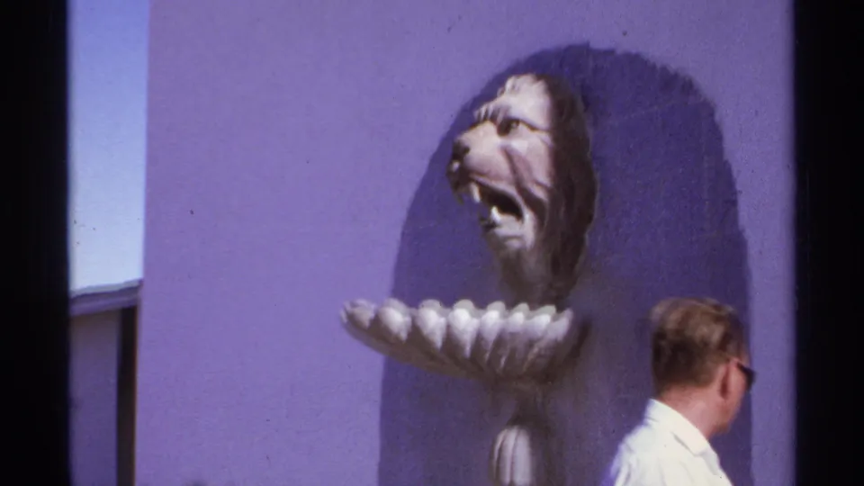 In Cabo San Lucas, Mexico in 1974, a man stands near a purple exterior wall with a roaring lion-head sculpture above him.