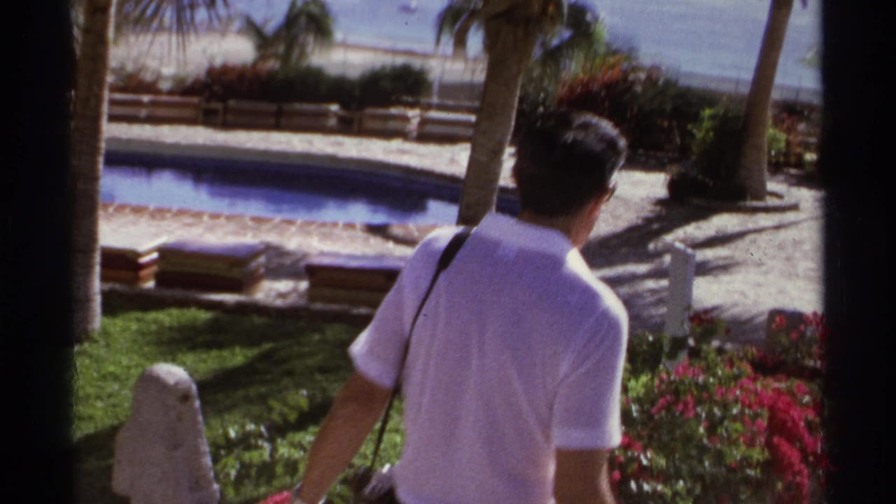 Man in a white polo shirt walking near a pool and palm trees.