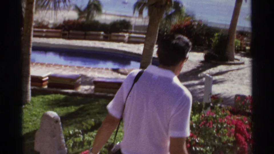 Cabo San Lucas 1974 frame showing a man in a white polo shirt walking through a tropical resort garden beside a pool.