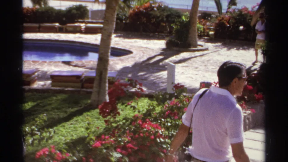 Cabo San Lucas 1974 home-movie frame with a man crossing a landscaped garden beside red flowers, grass, palms, and a curved pool.