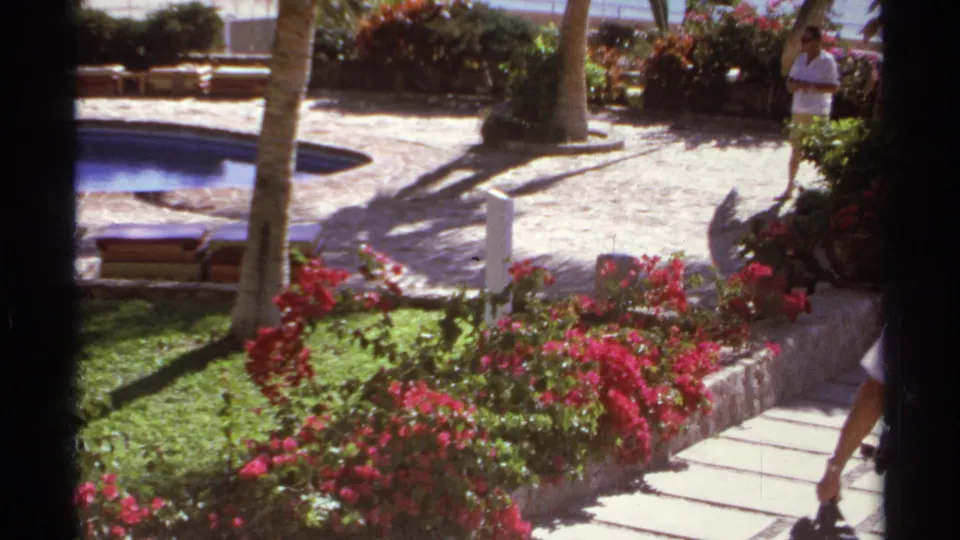Cabo San Lucas 1974 frame showing a resort garden path with red flowers, palm trees, pool edge, and a distant man in a white shirt.
