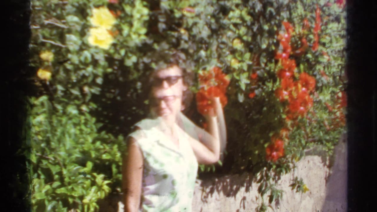 1974 Cabo San Lucas home-movie frame showing a woman in sunglasses near red hibiscus flowers and dense green foliage.