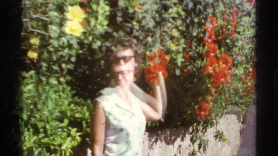1974 Cabo San Lucas home-movie frame showing a woman in sunglasses near red hibiscus flowers and dense green foliage.