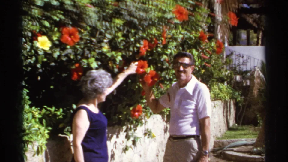 1974 Cabo San Lucas home-movie frame showing a man reaching toward hibiscus blooms as a woman stands nearby in the garden.