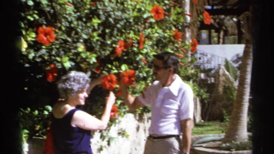 In 1974 Cabo San Lucas, a man and woman stand near red hibiscus flowers in a sunlit resort garden.