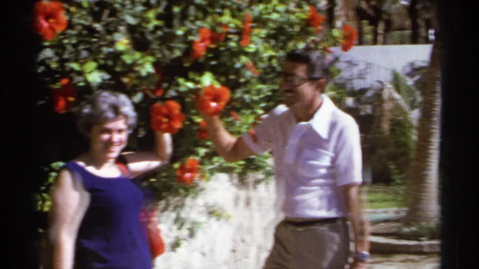 A 1974 vacation shot shows the woman and man framed closely by red hibiscus flowers and bright garden foliage.