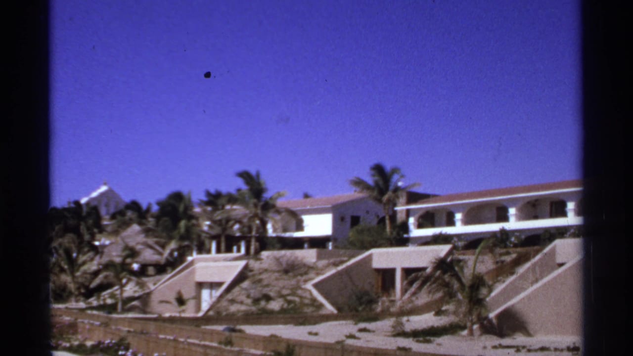 Terraced white resort buildings with arched balconies, palm trees, and sandy hillside under a blue sky.