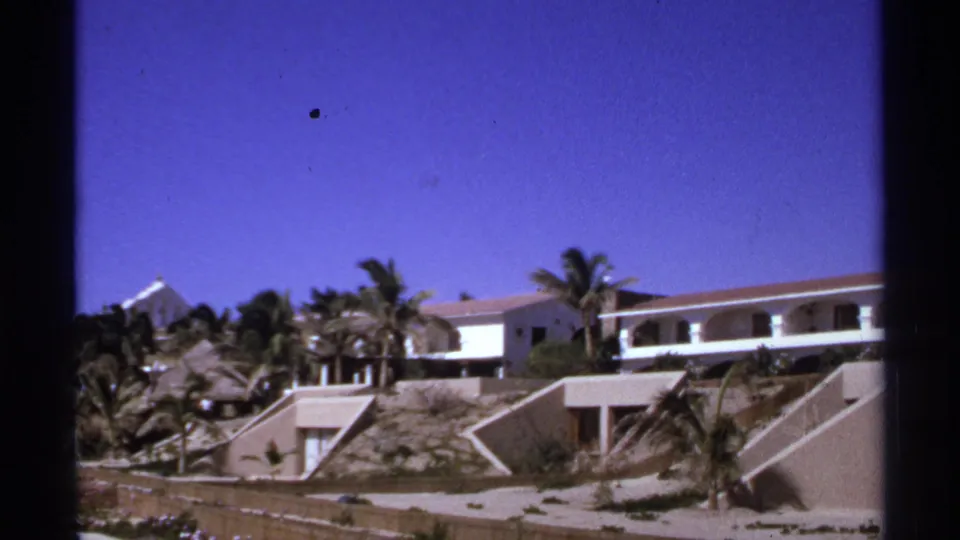 1974 Cabo San Lucas home-movie frame showing a modernist terraced resort with white balconies, palm trees, and sandy stepped architecture.