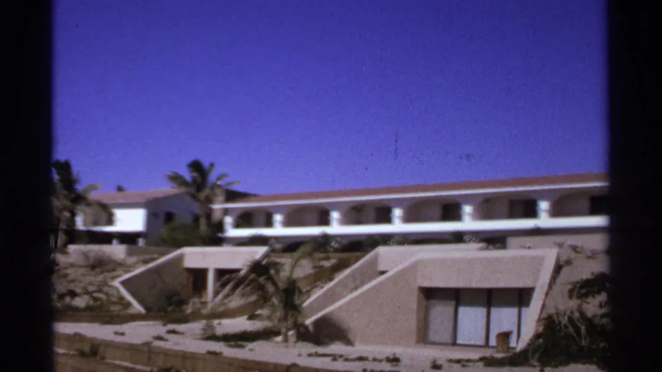 1974 Cabo San Lucas home-movie still showing terraced resort units below a white balcony building, with palms and saturated blue sky.