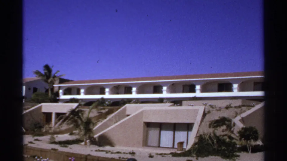 1974 Cabo San Lucas frame showing a modernist resort facade with arched balcony corridor, stepped walls, palms, and lower terrace doors.