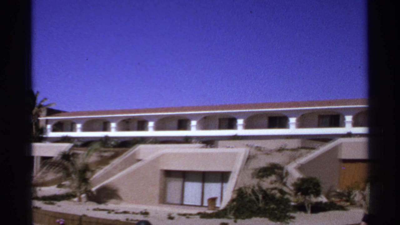 Modern resort building with white arched balcony, tan walls, hillside plants, and deep blue sky.