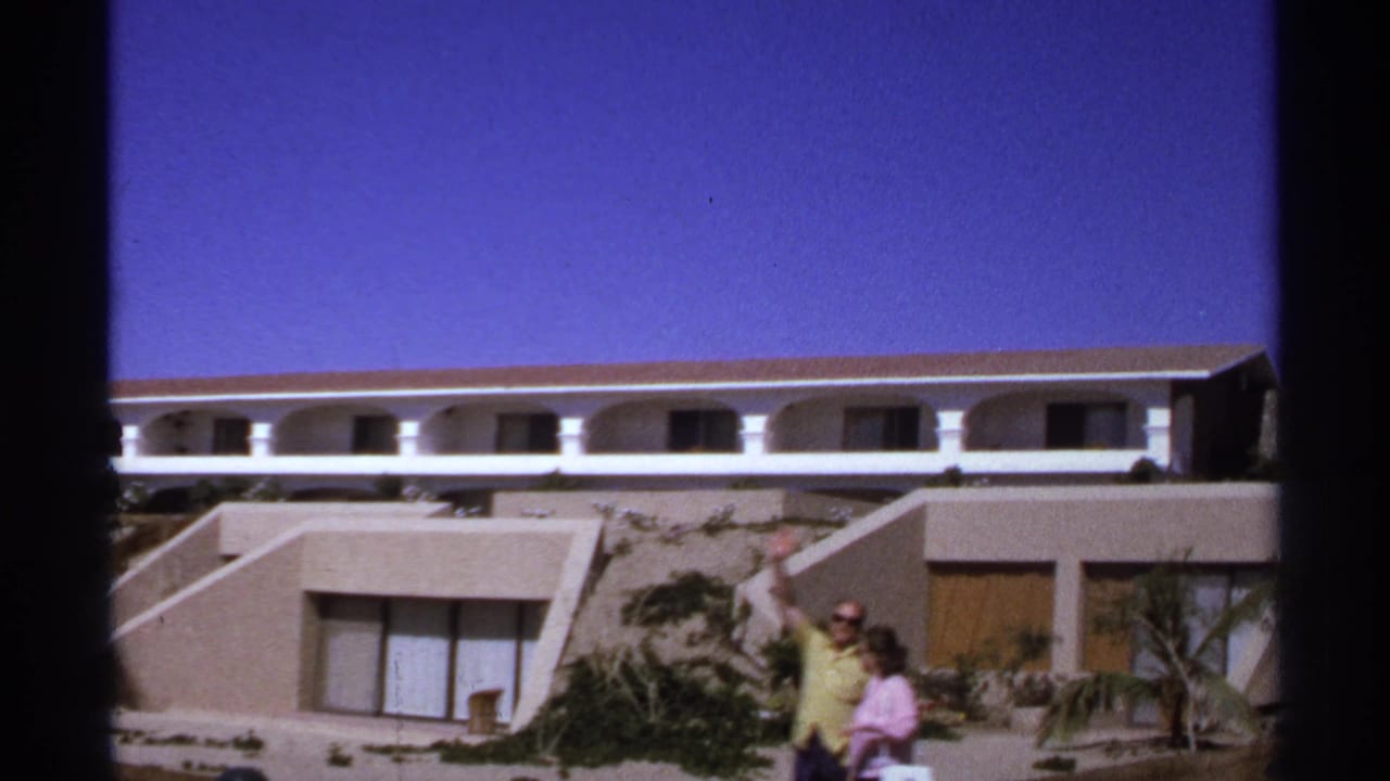 Two adults walk in front of a modern resort building as one waves toward the camera.