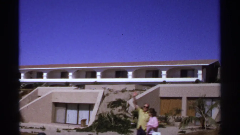 In Cabo San Lucas, two adults in bright 1970s clothing pass a hillside resort building while the man waves at the camera.