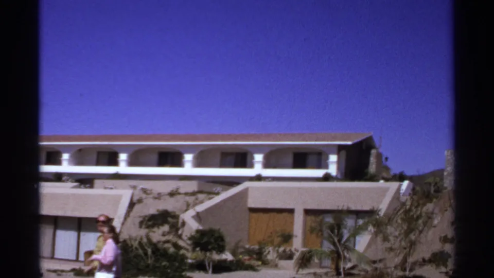 Two adults are partly visible near the edge of a 1974 Cabo San Lucas resort view, with tan walls and an arched balcony behind them.