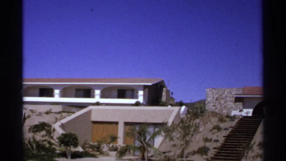 Cabo San Lucas resort exterior with arched balcony, tan stepped hillside, stone wall, and stairway under a clear blue sky.