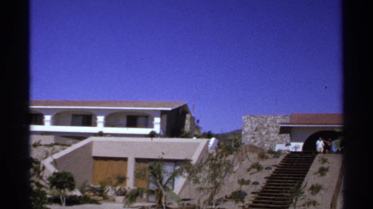 Hillside stone and stucco building with staircase, palms, and people near the entrance.