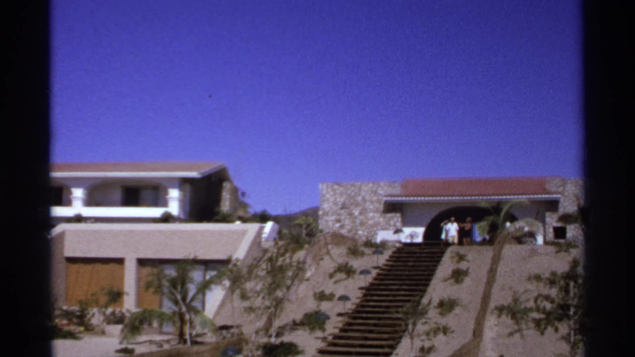 Stone hillside building with arched entry, staircase, small group of people, and bright sky.