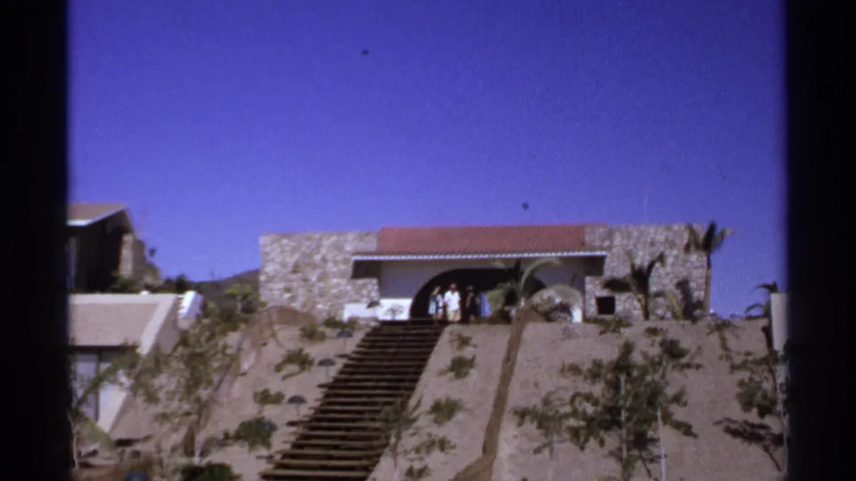 1974 Cabo San Lucas frame showing people at a hillside building entrance, with stairs, palms, stonework, and saturated blue sky.