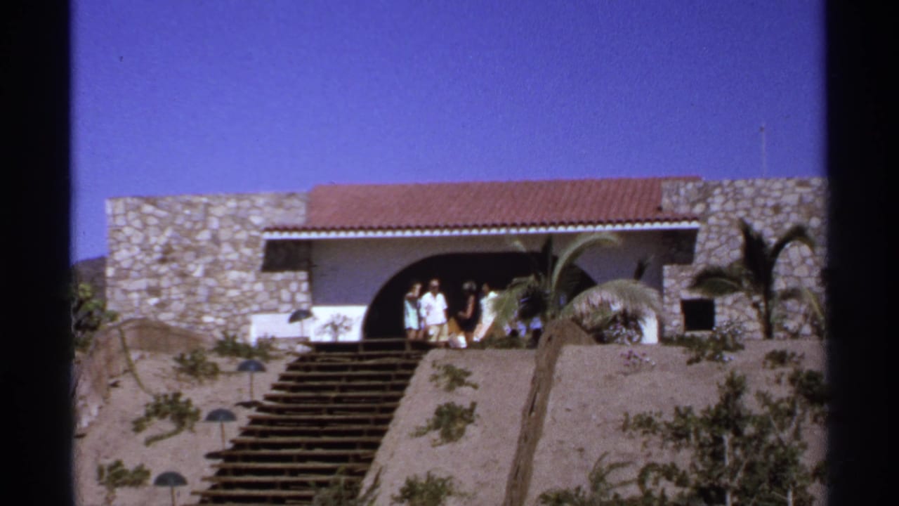 People in summer clothing stand by a stone house with a red tile roof and stairs.