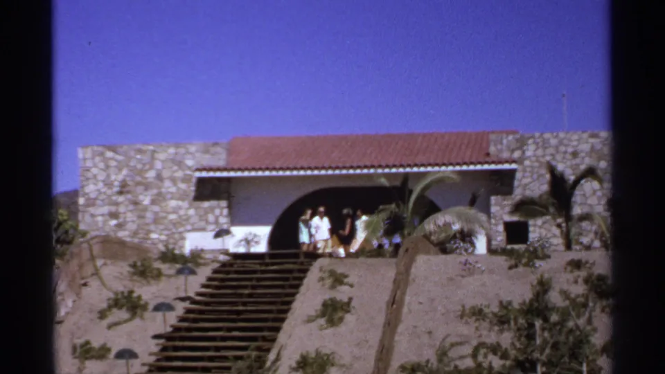 A group of adults in summer attire stands outside a stone and stucco hillside house in Cabo San Lucas, with steps and palms visible in the foreground.