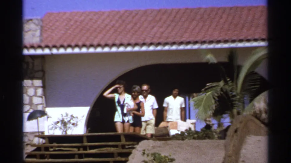 A closer 8mm frame shows adults in shorts and light clothing near the arched entry of a Cabo San Lucas hillside house, with palm fronds and a red tile roof above.