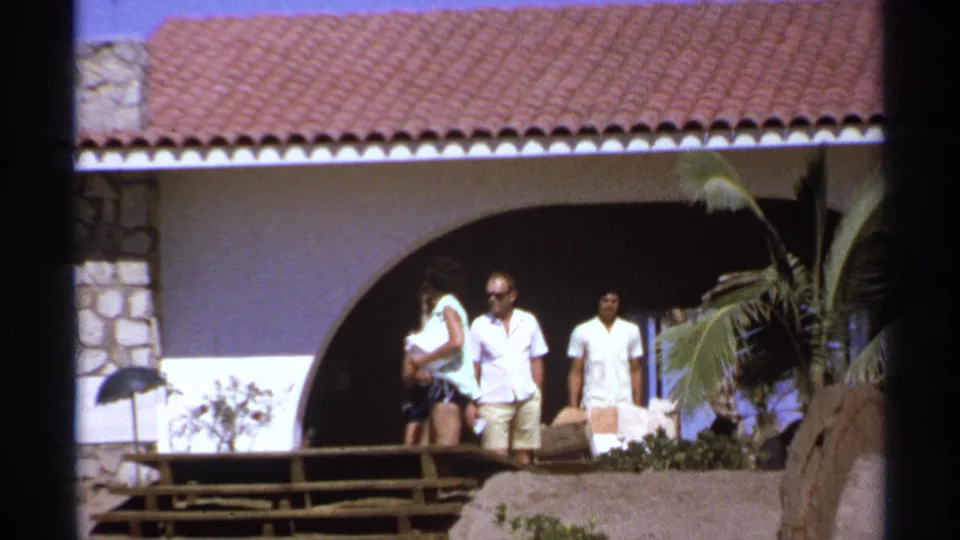 1974 Cabo San Lucas home-movie frame showing adults near a Spanish-style villa with a tiled roof, stone wall, deck, and palm fronds.