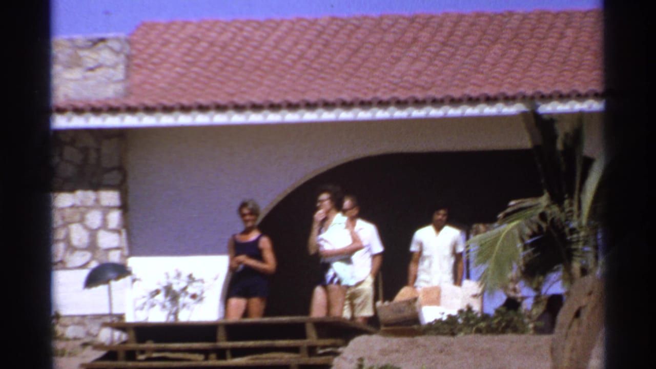 Four adults walk or stand outside a villa with arches, stonework, and palm fronds.