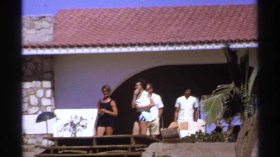 1974 Cabo San Lucas vacation frame with adults in swimwear and light clothing outside a Spanish-style villa and wooden deck.