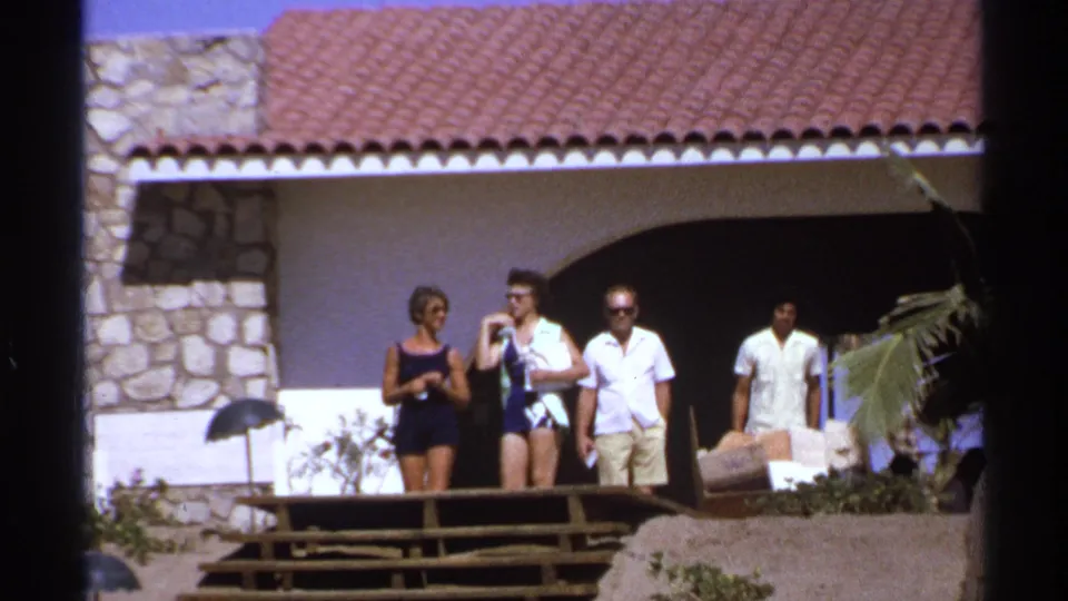 1974 Cabo San Lucas frame showing adults conversing on a deck outside a terracotta-roofed villa with stone details.