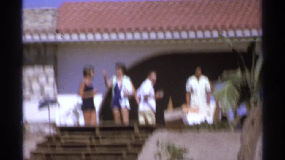 Soft, slightly blurred 1974 Cabo San Lucas frame with adults moving past a Spanish-style villa, deck, and palm foliage.