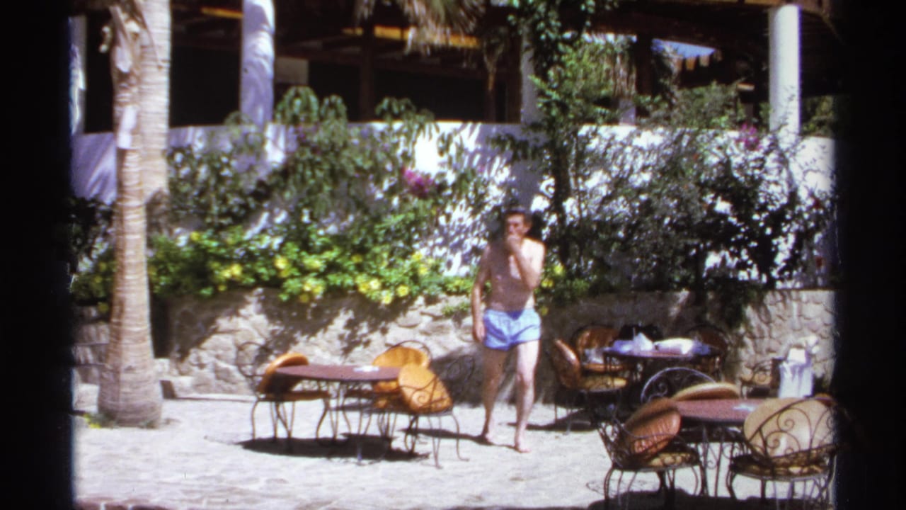 Shirtless man in blue swim trunks walking across a sunny courtyard with patio furniture.