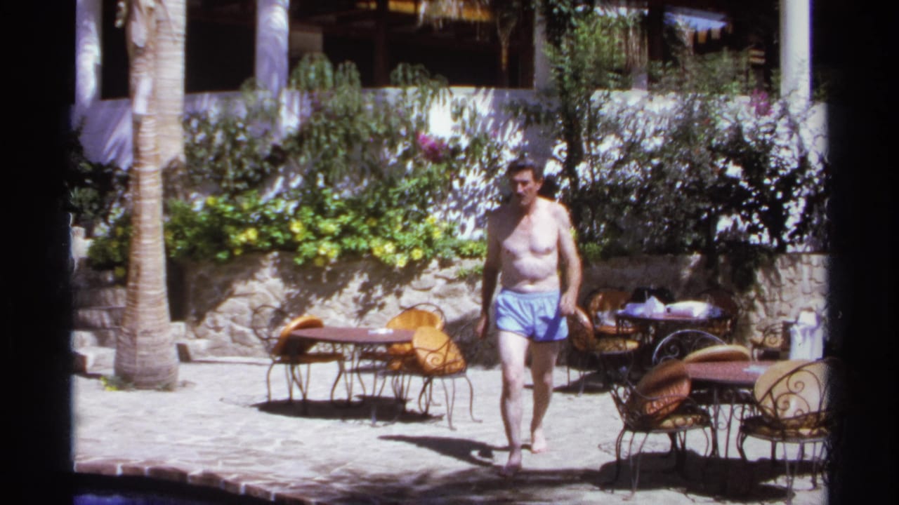 Shirtless man walking past outdoor tables and chairs in a bright courtyard.