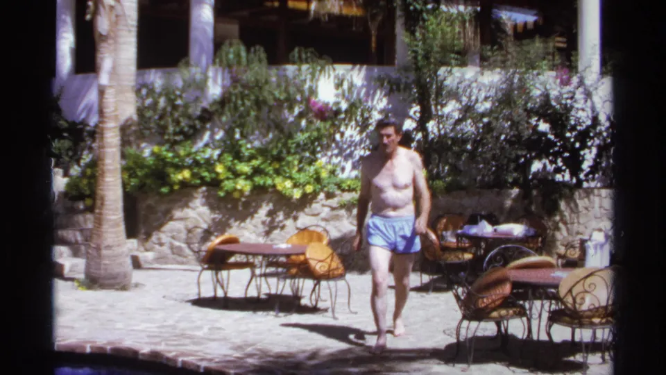 The man walks closer across the Cabo San Lucas courtyard, with palm trees, flowering greenery, and outdoor seating behind him.