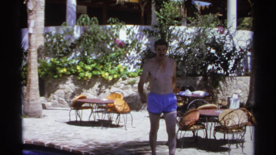 A shirtless adult man in blue trunks approaches the camera in a 1974 Cabo San Lucas courtyard framed by chairs, tables, palms, and shrubs.