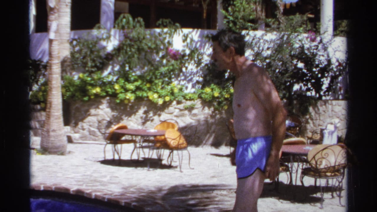 Man in blue swim trunks walks past a sunlit resort pool courtyard.