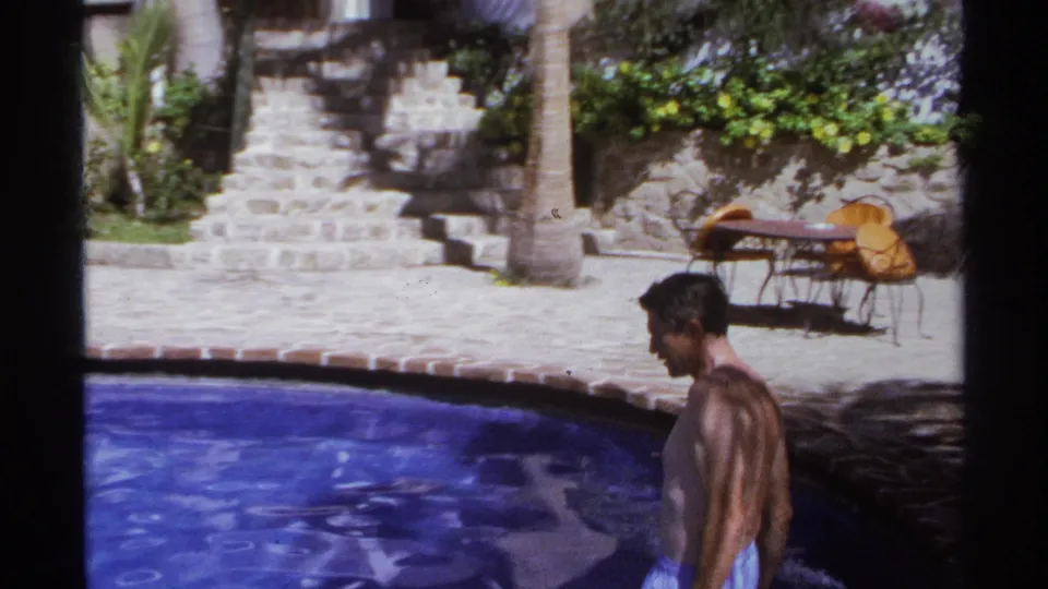 The man continues along the pool edge at the 1974 Cabo San Lucas resort, with stone steps, palm trunks, patio seating, and bright water in view.