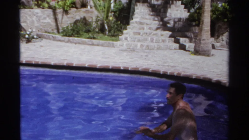 Adult man in blue swim trunks is partly submerged in the resort pool in Cabo San Lucas, with stone paving, stairs, palm trunks, and patio furniture around him.