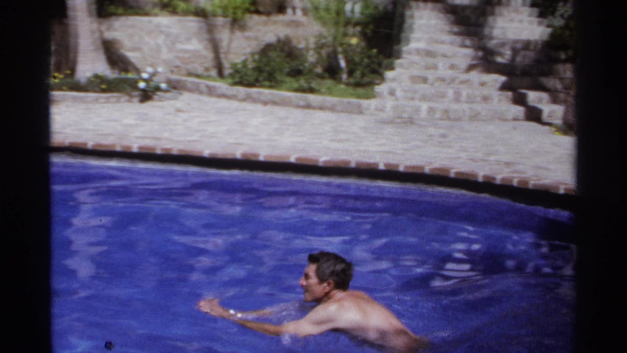 Two adult men swimming in a bright outdoor pool beside a stone patio.