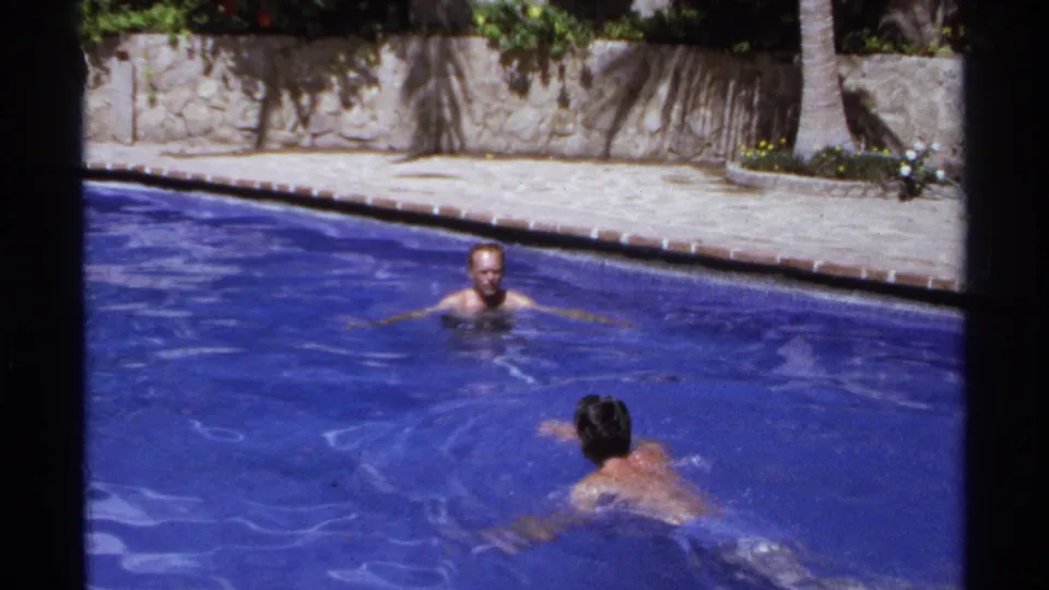 Two adult men appear to pause or swim toward each other in a sunlit Cabo San Lucas pool scene from 1974.