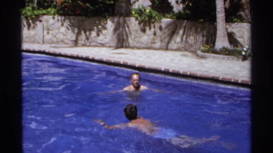 Two adult swimmers are seen in a bright blue pool at a Cabo San Lucas resort setting in 1974 home-movie footage.