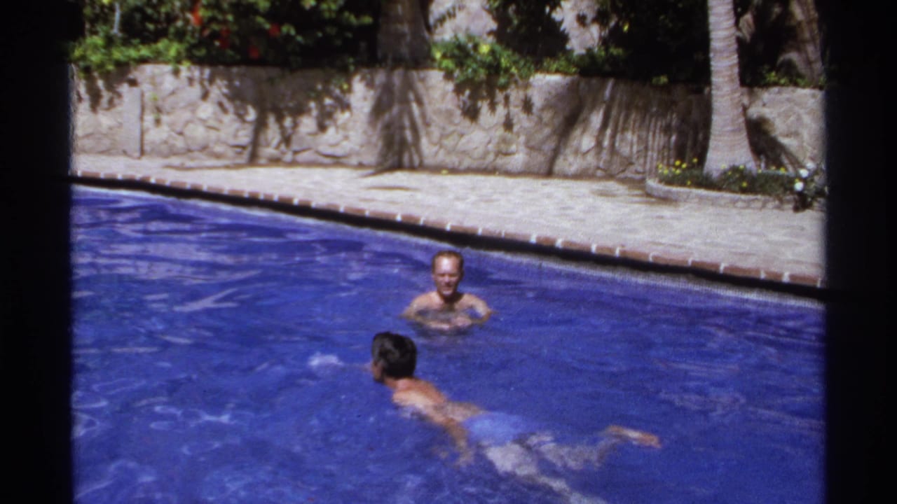 Two adult men in a blue outdoor pool, with palms and a stone wall behind them.