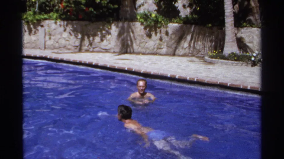 In 1974 Cabo San Lucas home-movie footage, two adult men swim and float in a sunlit blue resort pool near palms and a stone wall.