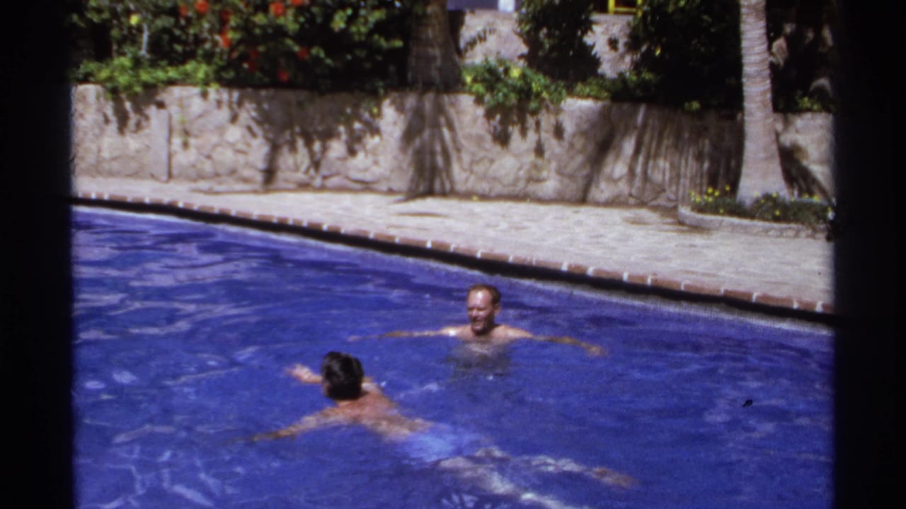 One man swims toward another man floating in a sunny outdoor pool.