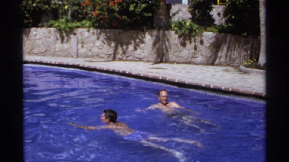Two adult men share a sunlit resort pool in Cabo San Lucas, with one swimming in the foreground and another floating near the pool edge.