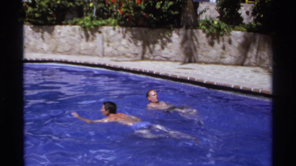 At the end of the clip, two adult men face each other in a bright blue resort pool, framed by palms, a stone wall, and 8mm film borders.