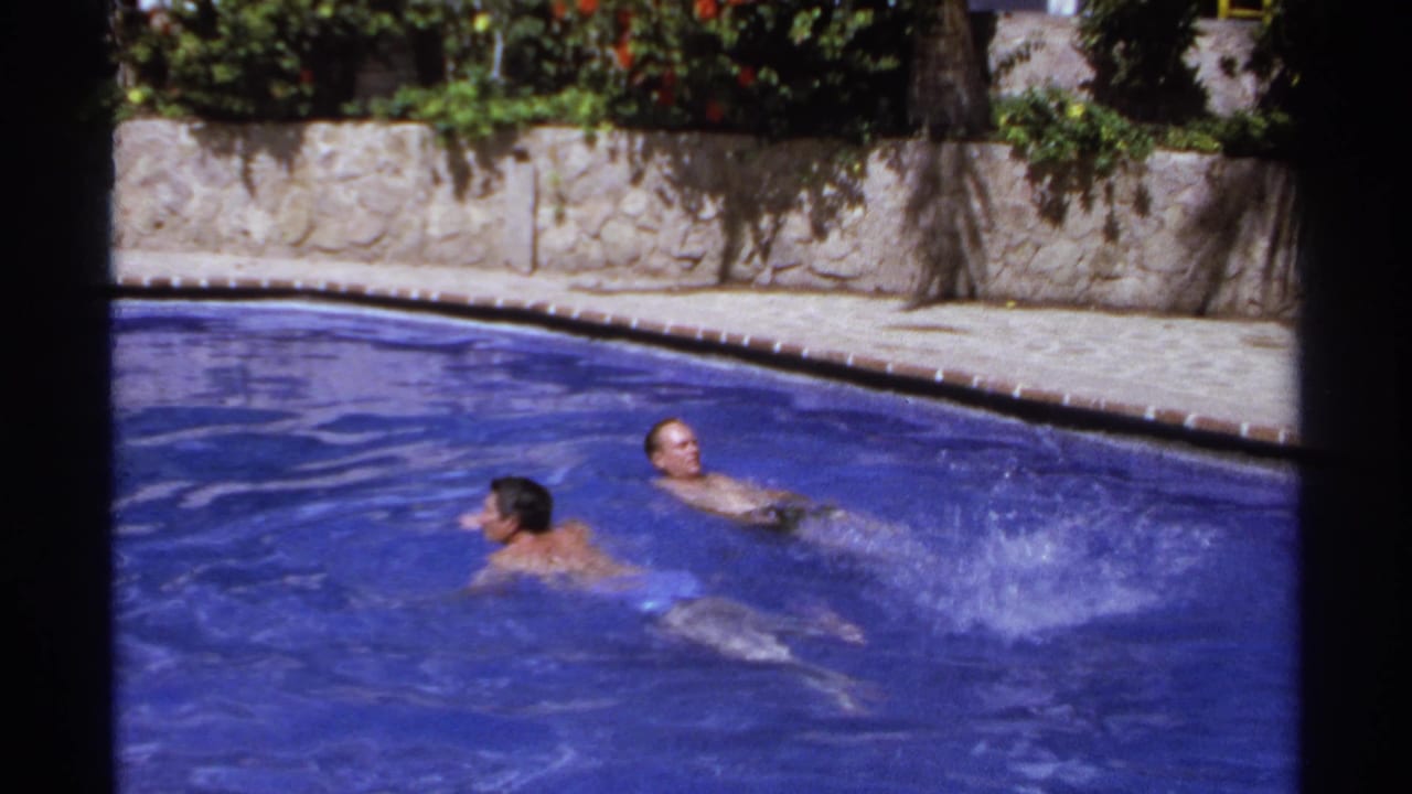 Two swimmers move across a blue outdoor pool with a stone wall and greenery behind them.