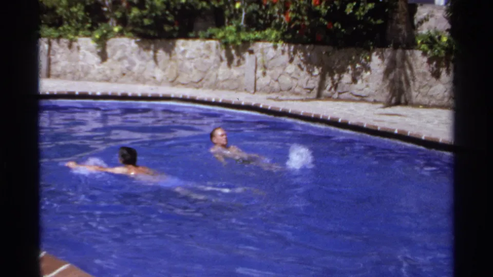 1974 home-movie pool frame with two young swimmers, a visible splash, blue water, and a stone garden wall in Cabo San Lucas.