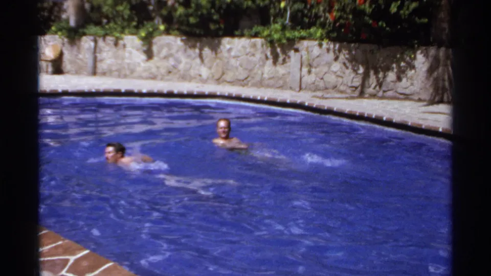 Two young swimmers are visible in a bright Cabo San Lucas pool scene from 1974, with faces partly seen and stone landscaping behind them.