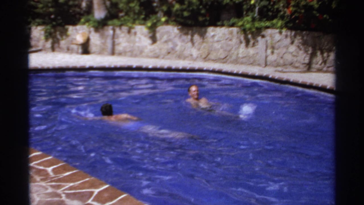 Two people swim in a blue outdoor pool beside stone walls and greenery.